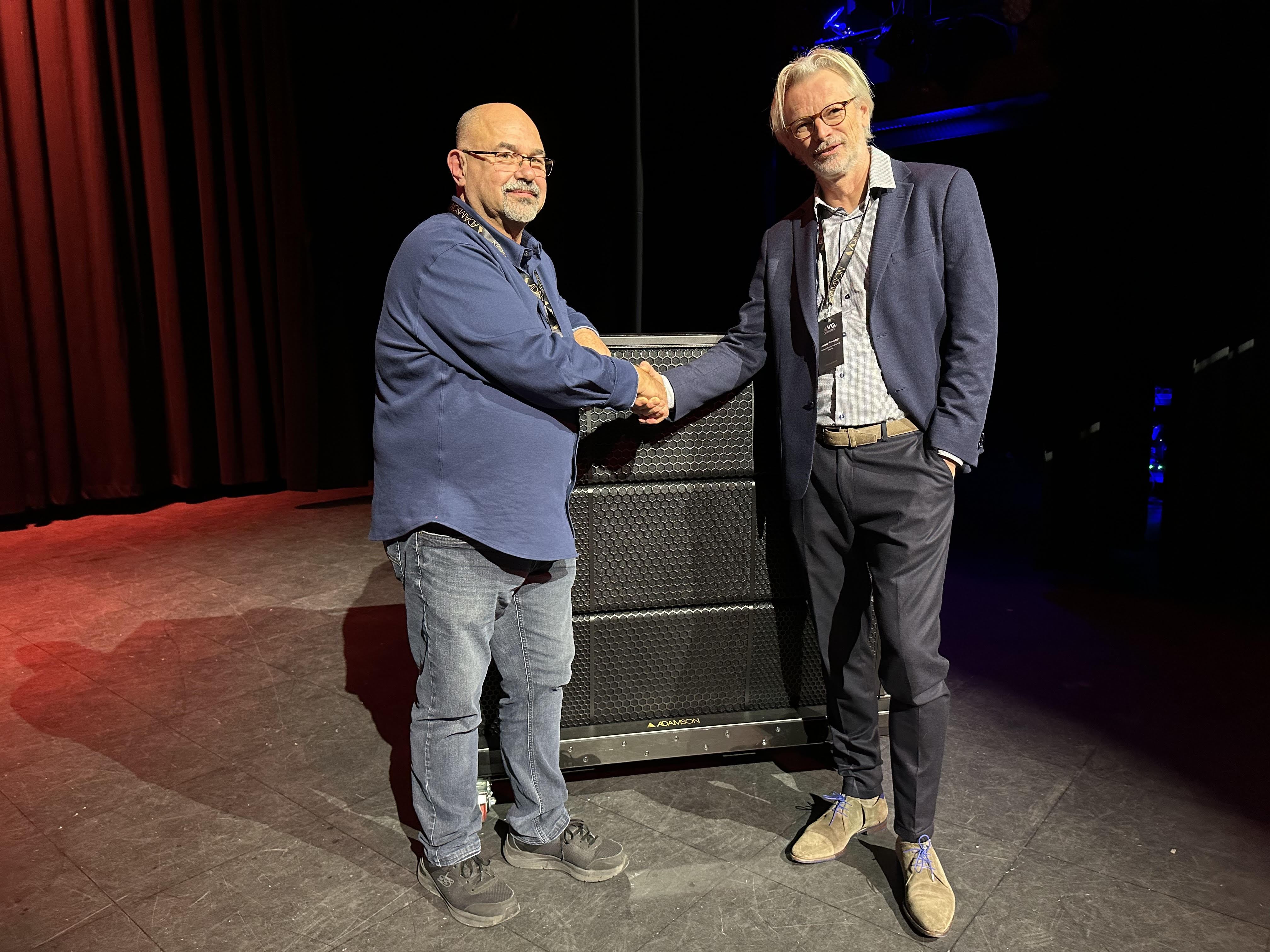 Vince Borrelli (left) and Jasper Ravesteijn (right) stand on a stage, shaking hands in front of an Adamson loudspeaker cabinet. The setting is dimly lit with stage curtains and theatrical lighting, highlighting their new partnership as Vince joins Adamson as Business Development Manager for the UK and Ireland.